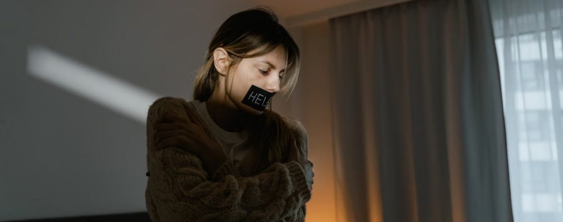 A young woman in a dim room wears a sweater and a help sign, expressing need.