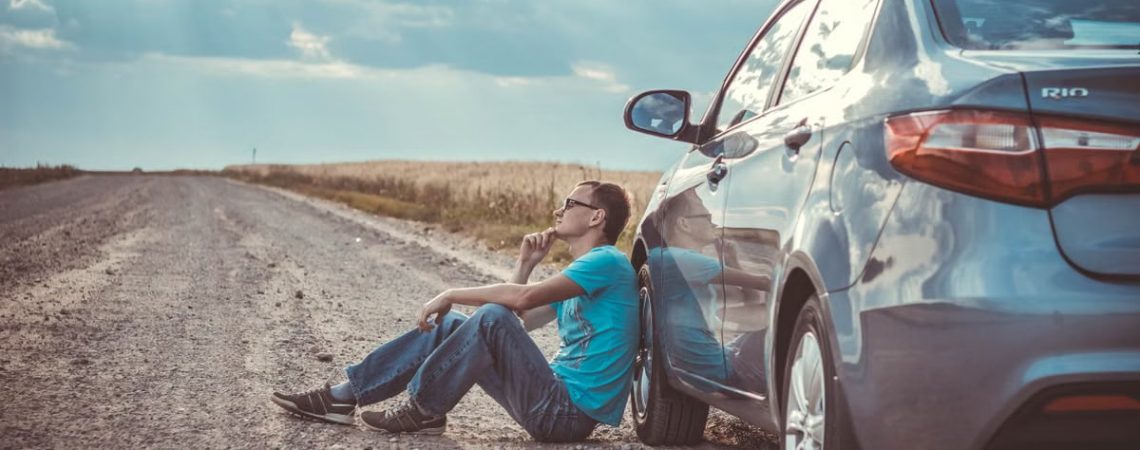 Man sitting by a car on a remote gravel road under a cloudy sky, deep in thought.